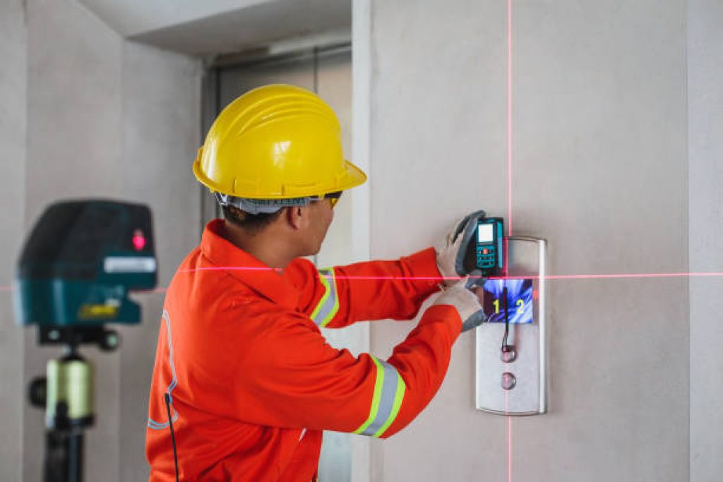 Asian Construction engineer in hardhat measuring wall with laser leveler next to elevator lift at construction site, Engineers at work checking building project with laser level machine during measurement work on site